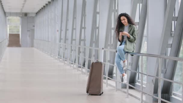 Ragazza viaggiatore passeggero donna siede su ringhiera nella stazione ferroviaria dell'aeroporto terminale con valigia bagagli guarda al telefono utilizza cellulare smartphone dispositivo mobile per cercare l'orario dei voli online — Video Stock