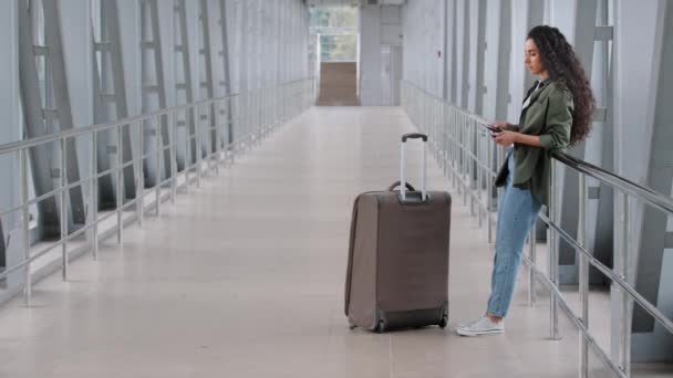 ヒスパニック系の若い女の子の旅行者女性の乗客は、空港ターミナルでスーツケースの荷物と一緒に立って飛行機のチケットを待っています到着時に電話画面で見て搭乗飛行旅行に行く — ストック動画
