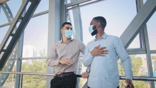 Два мультирасовых бизнесмена партнеры коллеги афро-американец и латиноамериканец менеджер босс стоять вместе в аэропорту с чемоданом носить защитные медицинские маски лица говорить разговор говорить — стоковое видео