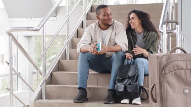 Pareja multiracial hombre africano y amigos de la mujer hispana sentados en los pies hablando en el aeropuerto cerca del tipo maleta celebración billete de avión chica con la comunicación de la mochila conversación antes de viaje — Vídeos de Stock