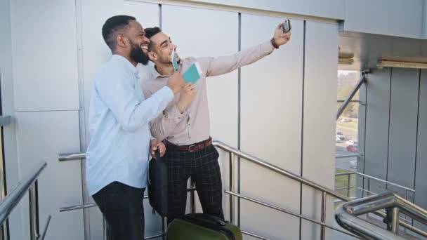 Dos amigos multirraciales socios de negocios feliz hombre hispano y chico africano sonriente con maleta de pie en el aeropuerto tomando foto selfie utilizando el teléfono móvil que muestra pasaportes billetes de avión videollamada — Vídeos de Stock