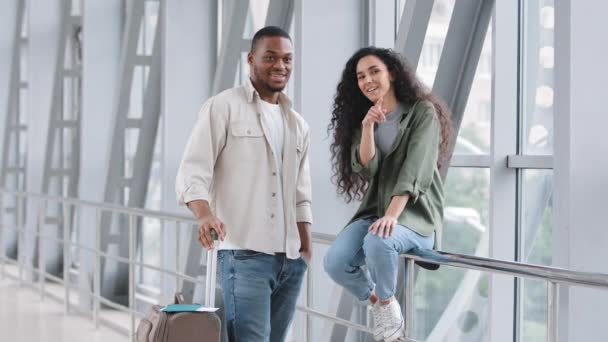 Couple multiracial hispanique femme et homme africain assis debout à l'aéroport avec valise parler conversation montrant la direction des mains en face d'eux parler avant l'embarquement Voyage de lune de miel — Video