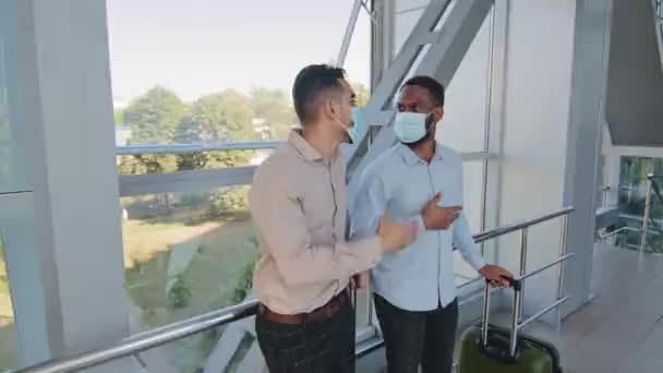 Dva multiraciální multi-etničtí přátelé obchodní partneři kolegové hispánský muž a africký šéf s kufříkem nosit obličejové masky stojící v terminálu letiště mluvit konverzace diskutovat o projektu — Stock video