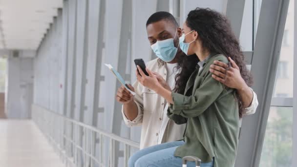 Wieloetniczna para nowożeńców Afrykański mężczyzna i Latynoska kobieta szczęśliwy małżonkowie nosić maski ochronne twarzy medyczne stoją na lotnisku z telefonu komórkowego rezerwacji hotelu w net oglądać lot harmonogram w aplikacji online — Wideo stockowe