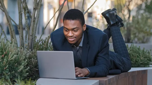 Усміхаючись впевнено задоволений молодий дорослий бізнесмен або корпоративний працівник, лежачи, використовує ноутбук, спілкується в соціальних мережах, готуючи промову. Змішана раса студентка чорного тисячоліття, яка навчається на відкритому повітрі — стокове фото