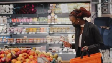 Tıbbi maskeli Afrikalı Amerikalı genç bir kadın süpermarkette elma meyvesi sebzesi seçiyor. Afro kız tüketici ürünleri almayı tercih ediyor. Taze yiyecek, market alışverişi.