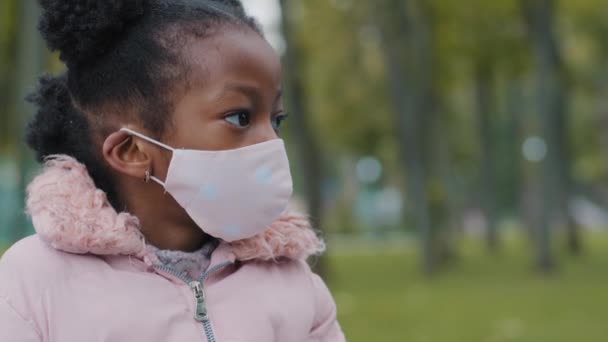 Close up kid face little girl in medical mask looks away turning head looking at camera in city street. Portrait of masked child baby pupil schoolgirl posing in park pandemic coronavirus quarantine — Stock Video