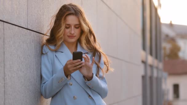 백인 사업가인 젊은 여성 이 핸드폰 온라인 쇼핑 앱을 들고 도시 밖에 서 있습니다. 스마트폰으로 카메라를 보며 현대 무선 기기에 대해 떠들고 있습니다. — 비디오