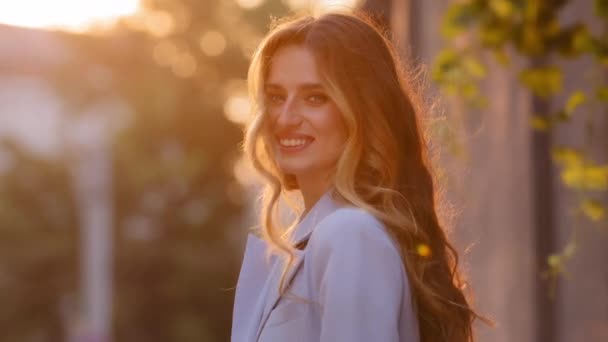 Portrét krásná atraktivní šťastná běloška s dlouhými vlasy stojí slunce paprsky pozadí při pohledu na kameru s úsměvem upřímně zubatý s bílými zuby. Millennial dívka pózuje na slunci s úsměvem — Stock video