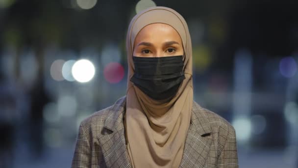 Fêmea islâmica close-up em máscara médica posando na cidade noturna. Mulher de negócios muçulmana vestindo hijab mascarado menina de pé olhando para câmera à noite fundo urbano ao ar livre pandemia coronavírus covid — Vídeo de Stock