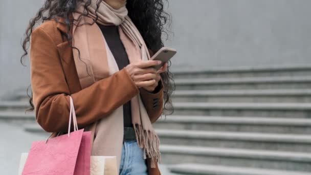 クローズアップ撮影認識できない若いスタイリッシュな女の子の買い物客の消費者の女性は、携帯電話オンラインショッピングアプリでチャット購入と明るいバッグを保持街の通りに立って — ストック動画