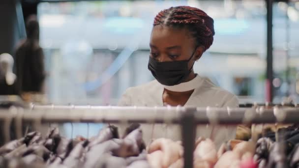 Afro americano mulher comprador Africano menina consumidor feminino comprador em preto máscara médica na loja de roupas desfrutando de venda descontos loja shopping escolher roupas corre em rack de vestidos comprar — Vídeo de Stock