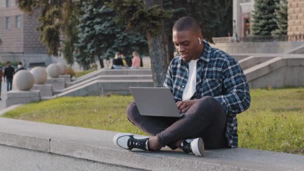 Afro amerikansk student kille afrikansk affärsman sitter utomhus i city street skriva på bärbar dator arbetar online med dator e-lärande lyssna på musik på hörlurar sång chatta i nätet — Stockvideo
