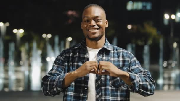 Afro americano hombre africano de pie en la noche ciudad cariñoso romántico retrato mirando cámara hacer forma de corazón con los dedos. Muchacho encantador mostrando amor bondad ternura apoyo signo con las manos — Vídeo de stock