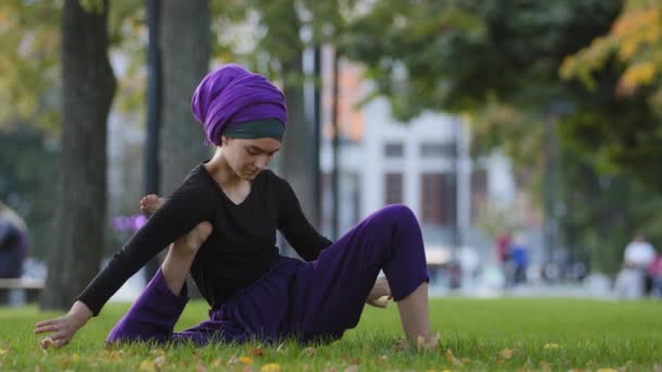 Κορίτσι σε hijab muslim γυναίκα σπορ κυρία θηλυκό γιόγκι κάθεται στο πάρκο της πόλης στο γρασίδι κρατά το πόδι κάτω από το χέρι κάνει τέντωμα λαβή με τα χέρια πρακτική γιόγκα προπόνηση τεντώνει σε εξωτερικούς χώρους αθλητικές ασκήσεις — Αρχείο Βίντεο