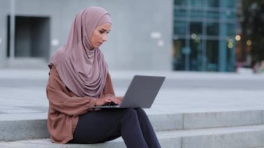Ciddi genç müslüman iş kadını, tesettürlü, bağımsız, Arap kız öğrenci, dizüstü bilgisayarlı, uzaktan kumandalı, uzaktan kumandalı, şehir daktilosuyla yazılmış, binanın yanında oturuyor.