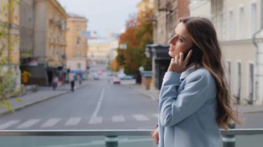 Mavi takım elbiseli başarılı genç bayan iş kadını balkonda cep telefonuyla konuşuyor. Kadın danışman, akıllı telefonla konuşur. Şehrin arka planında duran çevrimiçi konuşma cep telefonu alıcısı