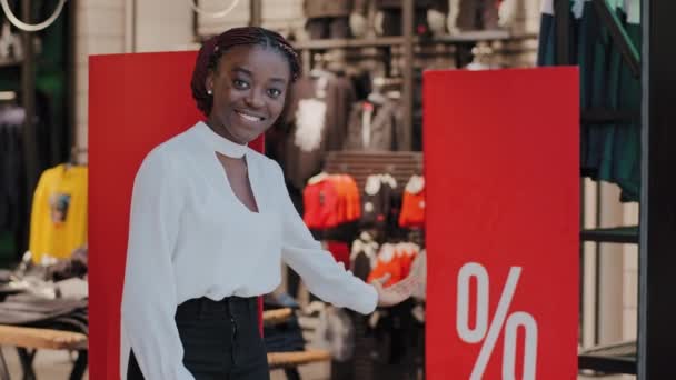 Kara Cuma konsepti. Gülümseyen Afro-Amerikan kadın satış elemanı Afrikalı kız danışman butik mağazası çalışanı alışveriş merkezinde duruyor büyük indirimli indirimlerden kıyafet almak için davet ediyor. — Stok video