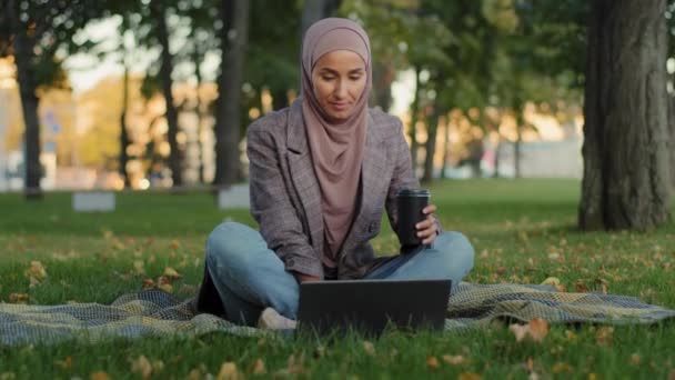 Glücklich lächelnde muslimische Geschäftsfrau Mädchen islamische Studentin trägt Hijab sitzt auf grünem Rasen im Stadtpark mit Laptop für entfernte E-Learning-Arbeit chatten online E-Mail checken mit Kaffee — Stockvideo