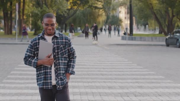 Pomyślny młody afro amerykański biznes człowiek facet afrykański student w okularach przeciwsłonecznych stoi w mieście ulica spacery przez ulicę ruchu drogowego samochód pieszy skrzyżowanie tło trzymając laptop uśmiechnięty idzie spacery — Wideo stockowe