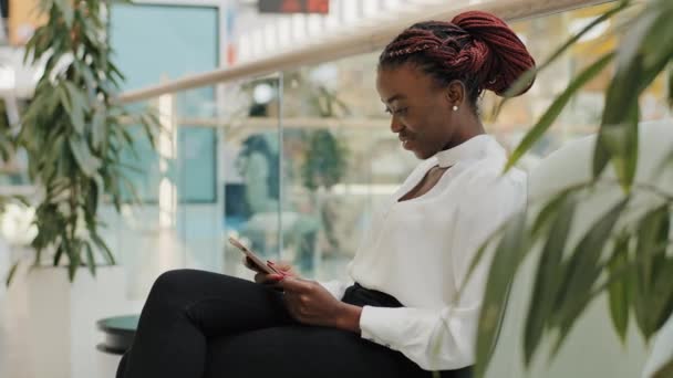 幸せなビジネス女性アフリカ系アメリカ人の女の子労働者のマネージャー若い女性が廊下オフィスに座っているスマートフォンを使用して携帯電話でチャットショッピングセンターで現代的な創造的な会社オンラインブラウジング — ストック動画