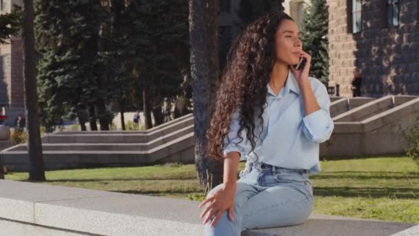 Atractiva chica joven feliz morena mujer hablando por teléfono móvil respuestas llamada sentado en la ciudad a la espera de la reunión espiando a alguien saludando de la mano invitando a venir llamando sonriente amigable — Vídeos de Stock