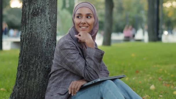 Leende drömmande kreativ pensiv flicka islamisk muslim affärskvinna kvinnlig författare journalist i hijab sitter på grön gräsmatta i parken kommer med nya roman skriva på laptop artikel inspiration — Stockvideo