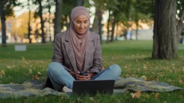 İslami Müslüman kız öğrenci internet öğretmeni iş kadını tesettüre girdi. Yeşil çimenlikte oturuyor, kahve içiyor, görüntülü sohbet konferansında bilgisayarla uzaktan konuşma yapıyor.