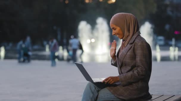 Vue de côté musulman islamic fille arabique entreprise femme étudiante professeur pigiste assis dans la nuit du soir fond de fontaine de la ville avec ordinateur portable faisant conférence vidéo en ligne parler saluant — Video