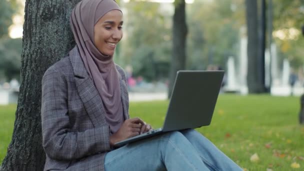 Happy usměvavý přátelský islámský obchod žena muslimský student dívka učitel nošení hidžáb sedí na trávníku v parku v blízkosti stromu mluví video hovor online chat konference mávání rukou pozdrav na notebooku — Stock video