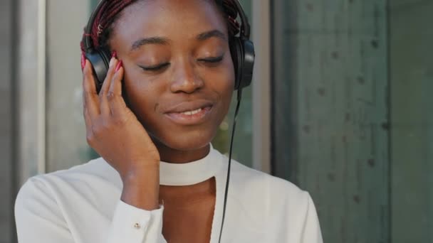 Hermosa chica afro-americana feliz despreocupada mujer africana joven mujer de raza mixta de pie en la ciudad escuchando música con auriculares auriculares audio en línea mp3 disfrutando de la canción cantar — Vídeos de Stock