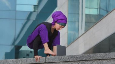 Genç güçlü iş kadını İslami Müslüman öğrenci dengede duruyor. Asana akrobatik hareketi dizüstü bilgisayarla yazı yazıyor. Şehir binası geçmişi bilgisayarın dinlenme işini bitiriyor. 