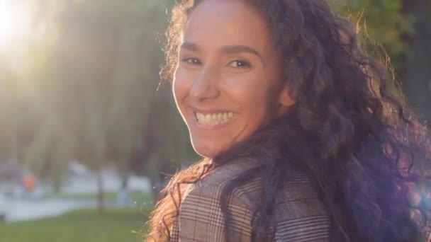 Smuk ung pige vender sig om ser på kameraet med tand smil poserende i sollys udendørs. Kvinde med langt sort krøllet hår vender i solstråler i parken. Attraktiv model med flyvende krøller – Stock-video