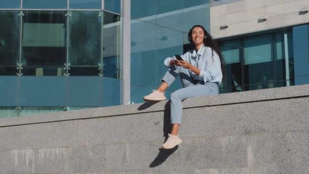 Szczęśliwy beztroski młoda brunetka dziewczyna kobieta pani siedzi na ulicy w mieście budynki miejskie tło słuchanie muzyki ze słuchawkami i telefonem komórkowym korzystających utwór dźwięk taniec z rąk — Wideo stockowe