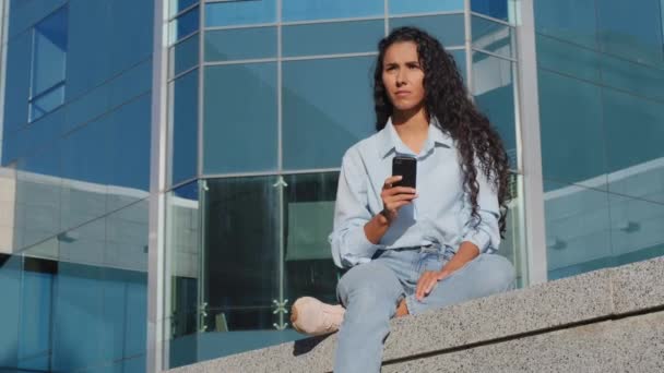 Pensivo estressado jovem menina com telefone celular senta-se na cidade perto do edifício pensa sobre o problema à procura de soluções se preocupa com dispositivo de bateria fraca mau dispositivo de comunicação celular smartphone — Vídeo de Stock