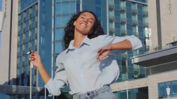 Kıvırcık saçlı, mavi tişörtlü güzel bir kadın sokak kenti arka planında dans ediyor eğleniyor zaferi kutlamaktan keyif alıyor. — Stok video