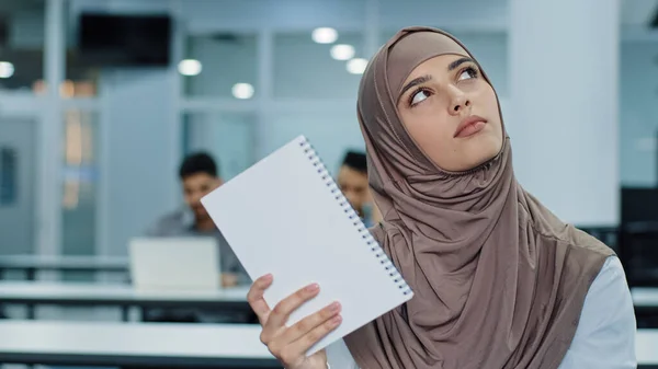 Pensiva unga arabiska kvinnliga arbetare i hijab sitter vid skrivbordet med hjälp av papper anteckningsblock gör anteckningar skapar nya idéer. Tankeväckande tusenårig blandras kvinna tittar åt sidan, funderar arbetsplanering — Stockfoto