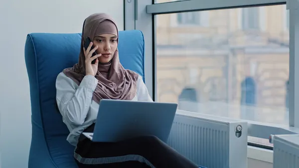 Pensiero arabo manager femminile in hijab seduto in poltrona confortevole parlare al telefono guardando monitor del computer portatile considerando l'idea, femminile pensieroso hanno conversazione cellulare pensare, prendere decisioni — Foto Stock