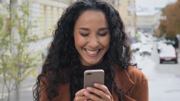 Close-up žena portrét krásná šťastná usměvavá bruneta žena s dlouhými kudrnaté vlasy stojí ve městě ulici při pohledu na mobilní telefon čtení dobré zprávy radosti chatování on-line v síti app e-shopping — Stock video