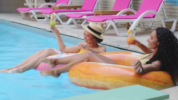 Красивые стройные девушки веселятся в летнем бассейне с холодными освежающими фруктовыми коктейлями. Взрослые женщины эмоционально празднуют праздник, смеются, брызгают водой, плавают на надувных спасательных буях — стоковое видео