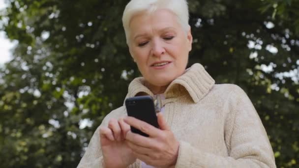 Χαμογελώντας ώριμη γυναίκα 60s γραπτών μηνυμάτων σε σύγχρονο smartphone gadget, ευτυχισμένη ηλικιωμένη γιαγιά 70s υπόλοιπο σε εξωτερικούς χώρους περιηγηθείτε στο διαδίκτυο στο κινητό τηλέφωνο, ελέγχει το ταχυδρομείο, καθημερινή εύκολη έννοια της τεχνολογίας χρήσης — Αρχείο Βίντεο