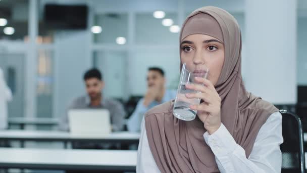 Assetata operaia araba indiana millenaria in hijab con bicchiere che beve acqua minerale pura in ufficio, pensosa impiegata donna disidratata che si gode l'acqua fresca. Concetto di stile di vita sano — Video Stock