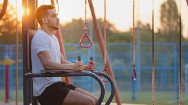 Yetişkin, yakışıklı, güçlü, sportif Hintli adam dizlerini kaldırıyor, sabah antrenmanı sırasında karın kaslarını çalıştırıyor. Orta Doğulu atletik genç adam spor sahasında egzersiz yapıyor, aletleri kullanarak kaslarını şişiriyor.
