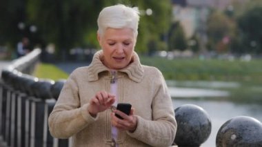 Modern akıllı telefon zımbırtısıyla mesajlaşan olgun bir kadın beklenmedik haberler okuyor, şaşırıyor, anneanne cep telefonunda internete bakıyor, zorluk yaşıyor, uygulama kullanarak hata yapıyor, internetten sipariş veriyor.