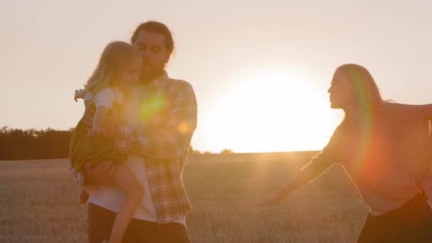Щасливі сімейні вихідні в літній вечір на фоні сонця пшеничного поля. Догляд за бородатим батьком тримає маленьку доньку молоду веселу матір, яка грає з дитиною, вдаючи, що літак грає весело — стокове відео