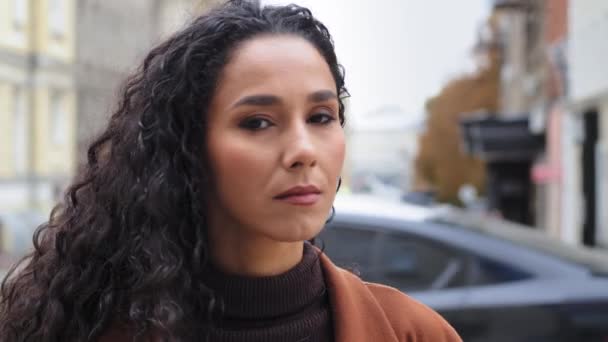 Close-up pensive serious sad millennial girl 30s woman brunette hispanic lady female with long curly hair and natural make-up looks to side turns head looking at camera standing in city windy weather — Stock Video