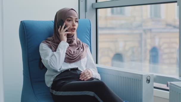 Sonriente Relajada árabe gerente milenaria en hijab sentado en sillón cómodo hablando por teléfono, consultoría, hacer llamadas de negocios en el trabajo, joven trabajadora o cliente hablando por móvil — Vídeos de Stock