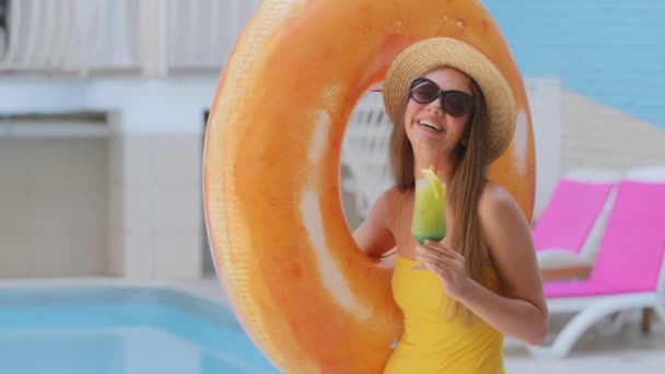 Mulher loira branca bronzeada bonita e esbelta em óculos de sol bebe suco de frutas por piscina no hotel spa. Menina europeia em maiô desfrutar de férias de luxo com coquetel, carrega lifebuoy anel inflável — Vídeo de Stock