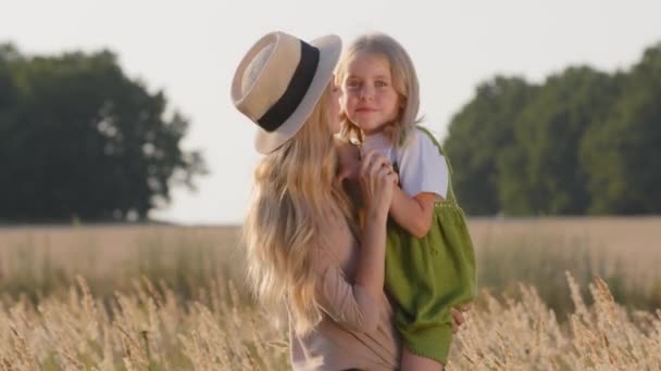 Genç, güzel anne, hasır şapkalı sarışın kadın buğday tarlasında duruyor. Küçük kız gülüyor, çocukla konuşuyor. Doğada birlikte vakit geçirip eğleniyorlar. — Stok video