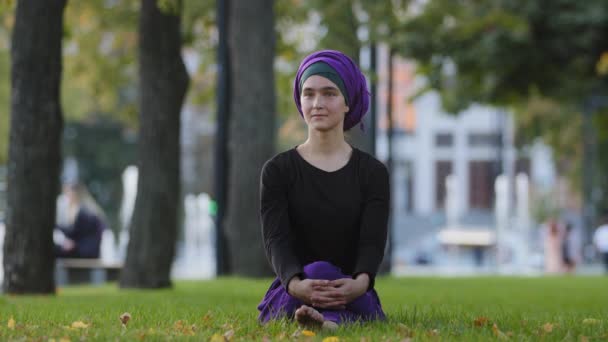 Beautiful muslim young islamic girl in purple hijab female buddhist sits on green grass lawn in city park outdoors enjoying good summer weather day sitting in nature dreaming thinking contemplating — Stock Video